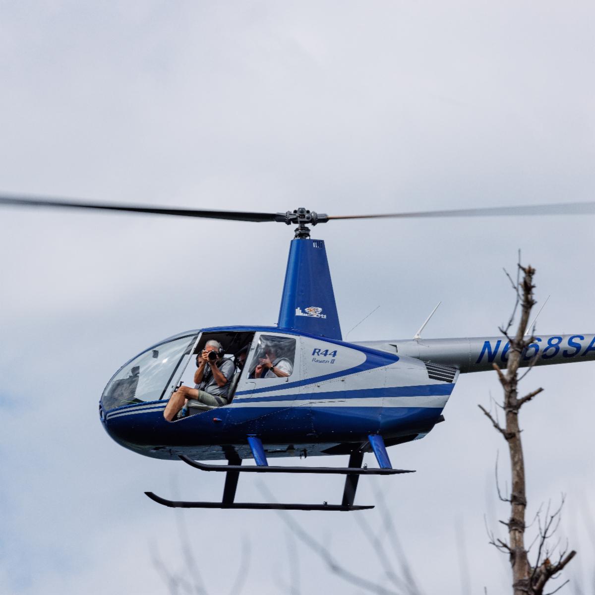 A silver and blue helicopter hovers in the air. A man holding a camera can be seen through one of the doors.
