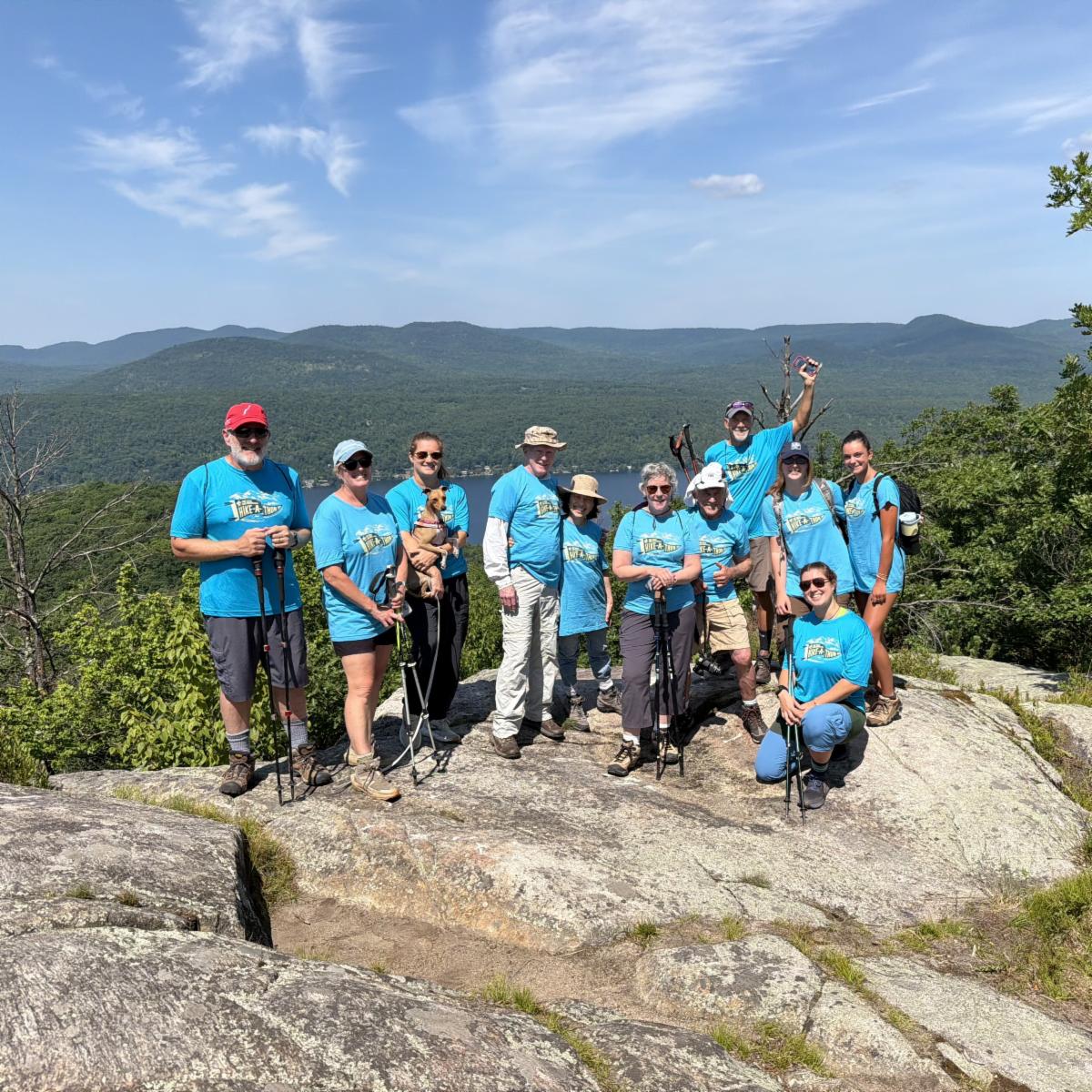 A group of a dozen people of various ages wearing blue shirts stand together on top of a mountain. 