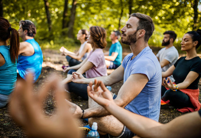 A group of people sitting down with their legs crossed, and each hand facing the sky with their thumb and index finger touching eachtoher