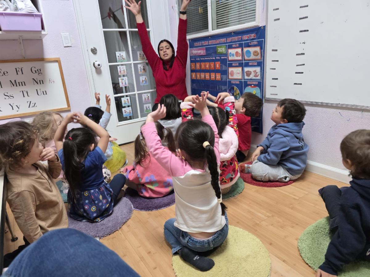 A teacher with raised hands sits with a group of children during a group activity