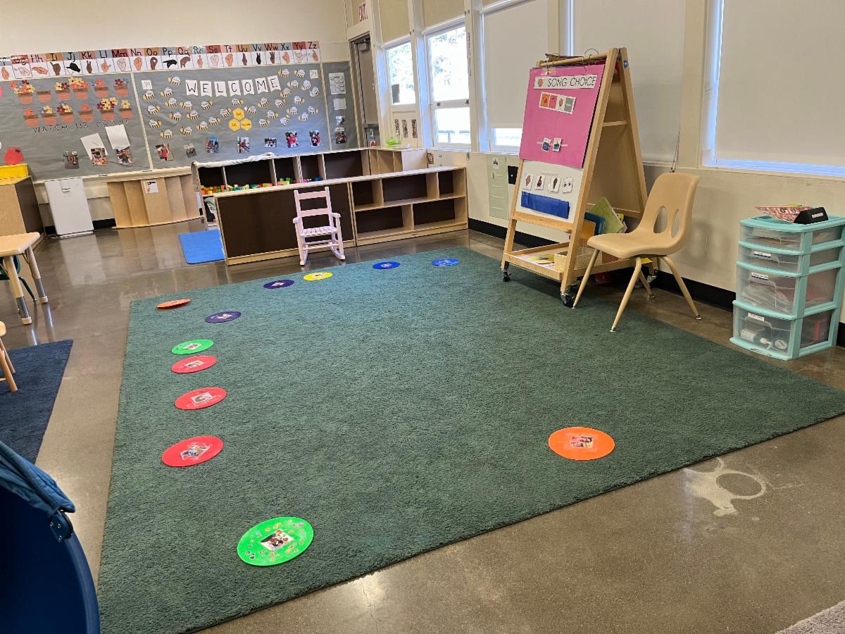 A green carpet and floor dots in the foreground with an easel and shelving in a preschool classroom