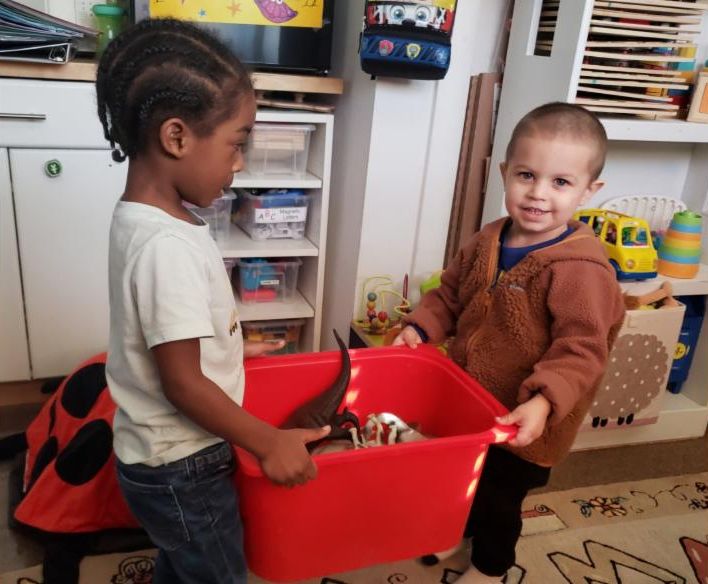 Two children carry a red container full of toys and materials together