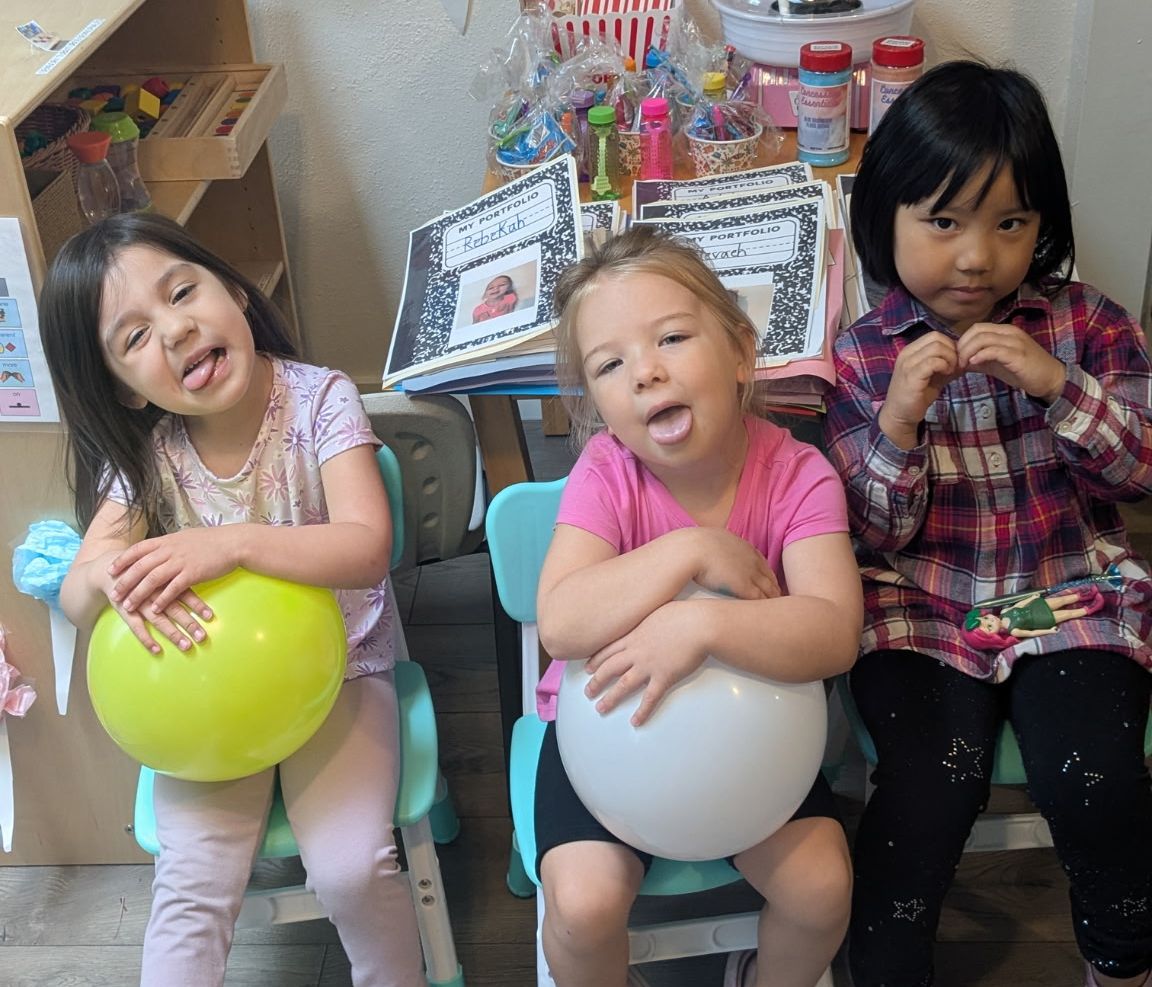 Three children sit on chairs while two hold a balloon and make silly faces