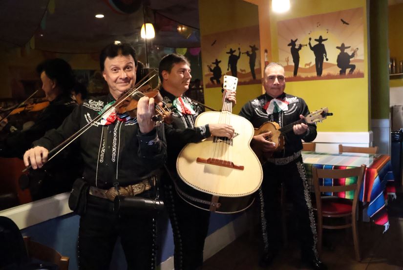 A mariachi band performs inside a restaurant. Three musicians in traditional black mariachi outfits play the violin, guitar, and vihuela in front of a festive mural.