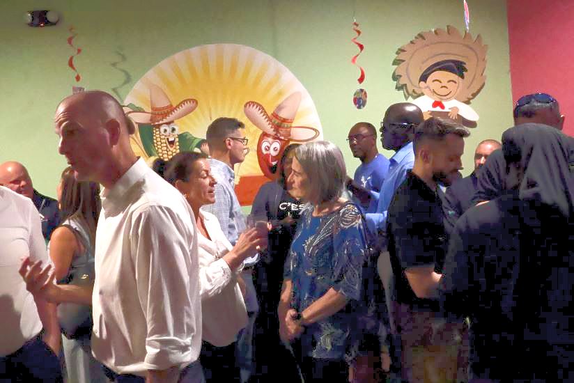 A group of people mingles inside a restaurant decorated with cartoon-style sombrero characters on the wall. The setting appears festive, with hanging red streamers and a warm ambiance.