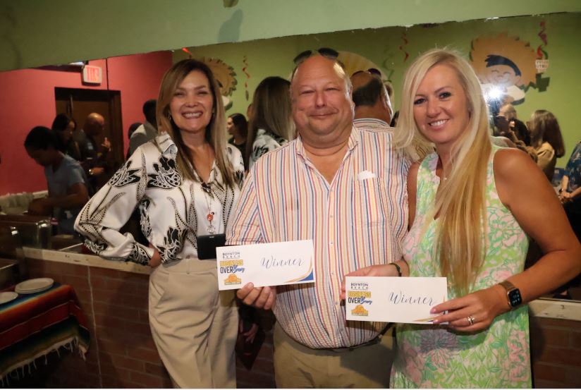 Two women and a man pose for a photo, smiling and holding "Winner" certificates from a Boynton Beach Business Over Brews event. A colorful restaurant backdrop and reflection in a mirror are visible behind them.