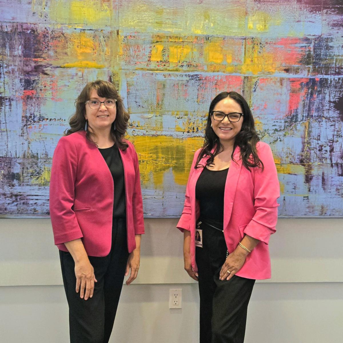 Photo of two women standing in front of a large modern looking colorful painting. Both women are wearing glasses and wearing similar outfits with hot pink blazers and black underneath. They are smiling and posing for the photo together. 