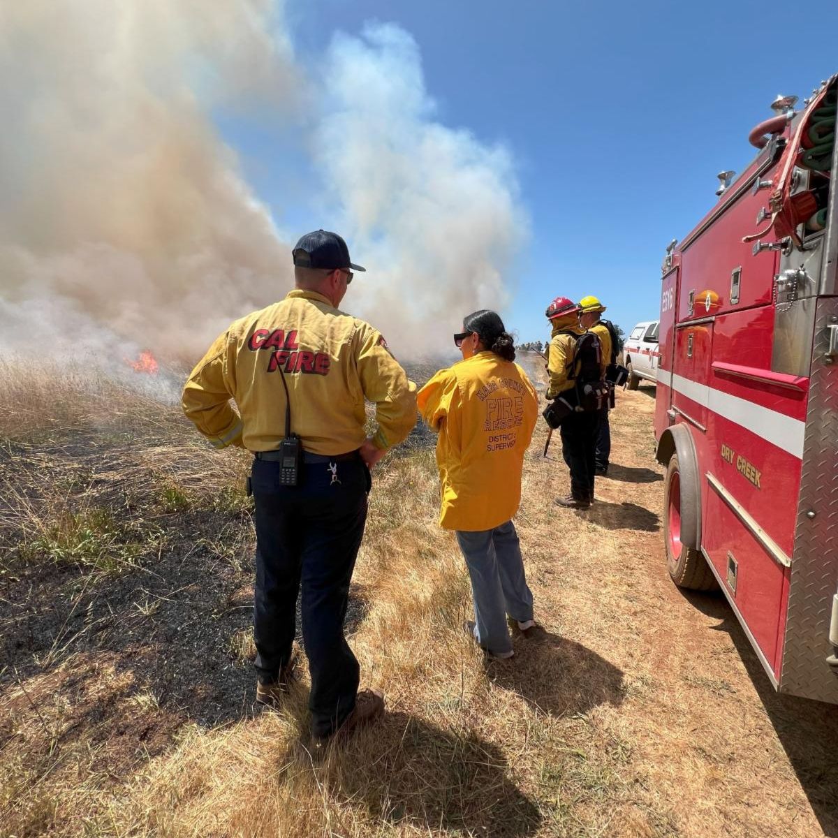 Photo of two adults standing with their backs to the camera, one male and one female. The male has a baseball cap on and the back of his jacket reads 'CALFIRE'. The back of the womans jacket reads 'Napa County Fire Rescue District 5 Supervisor'.A couple other adults in firefighting gear are in the foreground. They are all standing next to a fire engine on the right. To the left is burnt ground and fire and smoke in the foreground on the left. 