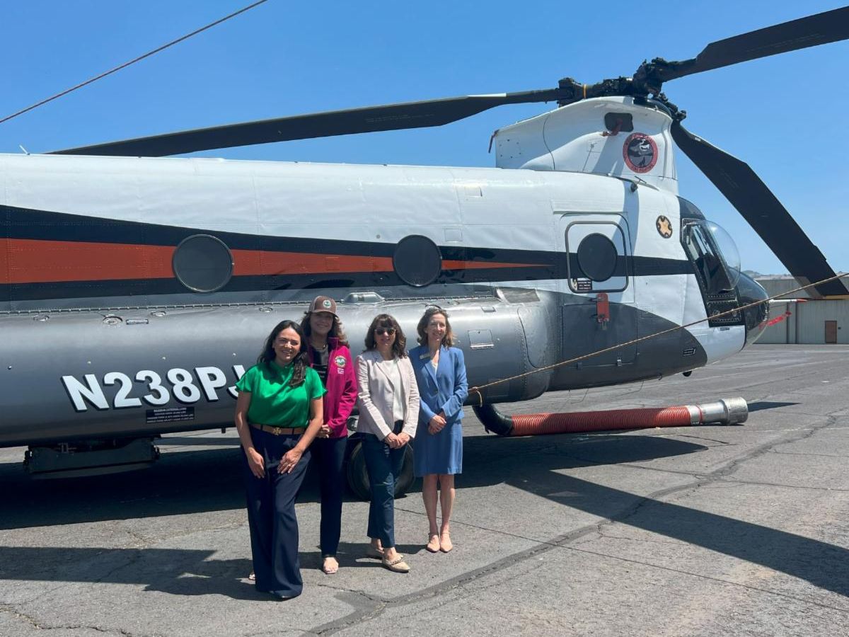 Photo of four Napa County Supervisors standing in front of a helicopter. The four women stand in front of a large white and gray helicopter with black and red accents on a sunny day. The helicopter's tail number N238P is visible on the side and its large rotor blades extend overhead. The group is standing on a paved surface with visible cracks and the background includes a clear blue sky.
