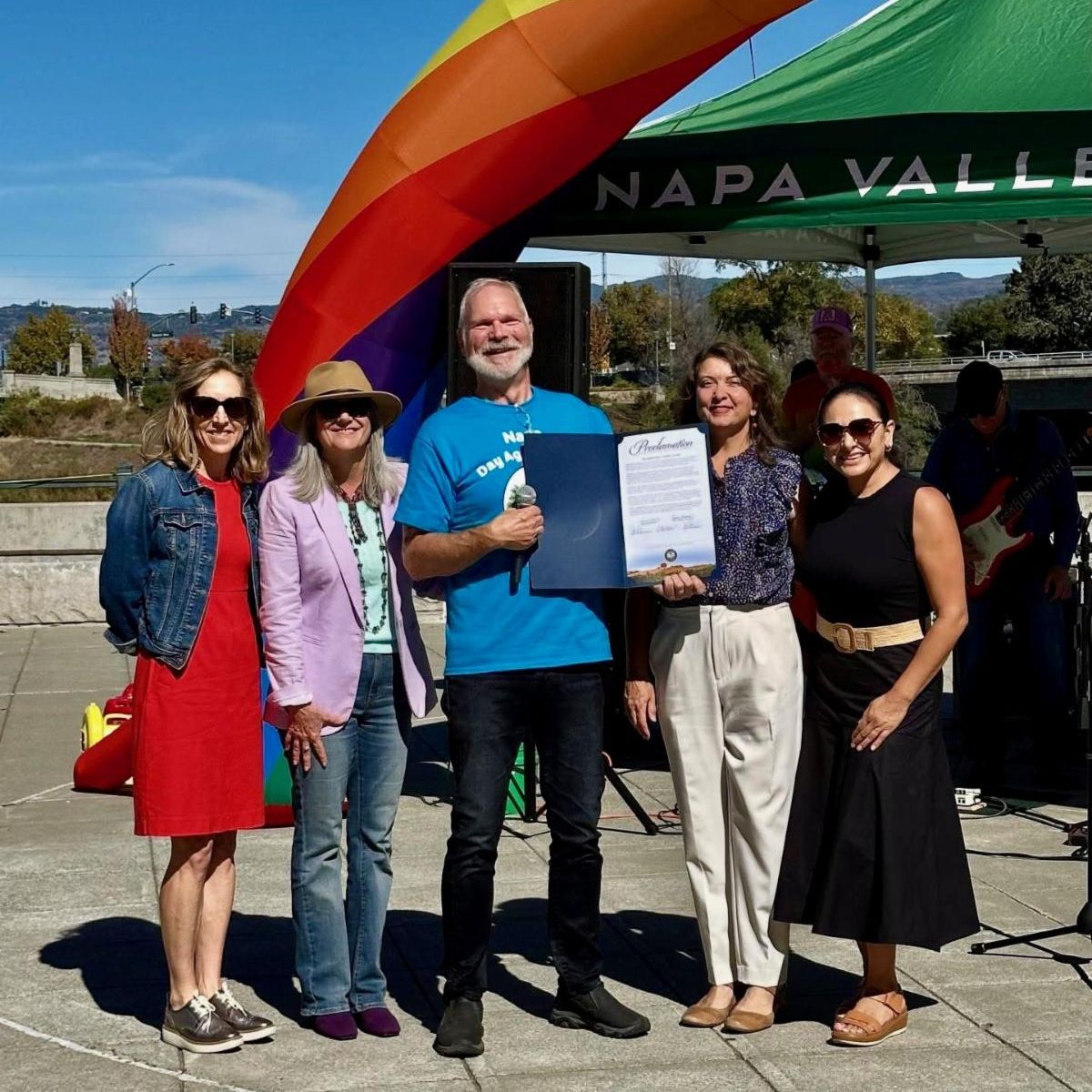 Photo is of a group of 5 people standing together posing for a photo. A large rainbow arch can be seen behind them. In the middle a man with a white beard is holding a proclamation for Napa Day Against Hate. On either side of him are two women. The women are four members of the Board of Supervisors including Anne Cottrell. Joelle Gallagher. Amber Manfree. And Belia Ramos. 