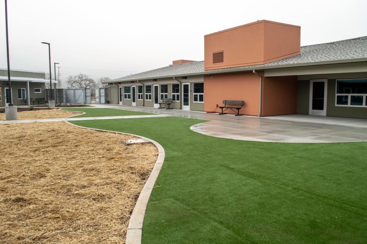 Photo of an outdoor courtyard area of a modern behavioral health treatment center. The scene features a curved concrete walkway bordered by green artificial turf and a section of straw covered ground possibly for future landscaping. The buildings are single story with a mix of earth tone colors including olive green walls and an orange accent structure. Several benches are placed along the walkway and the area appears clean quiet and secure with a tall metal fence and gate in the background. The overall environment looks calm and structured designed for therapeutic and recreational use.