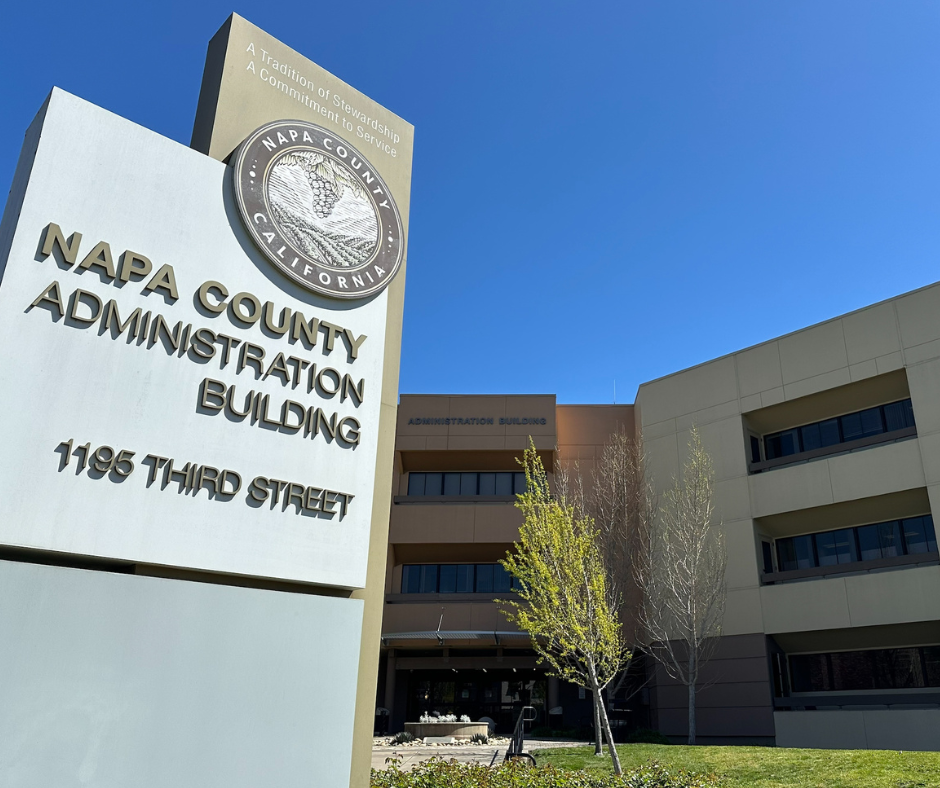Photo of a daytime view of the Napa County Administrative Building located at 1195 Third Street in Napa California. In the foreground a large sign displays the Napa County seal and the building name and address in raised letters. The building behind the sign is a modern three story structure with beige and brown tones large windows and a clean well maintained exterior. Youn trees with early spring leaves and trimmed landscaping are visible in front of the building under a clear blue sky. The inscription at the top of the sign reads A Tradition of Stewardship A Commitment to Service.