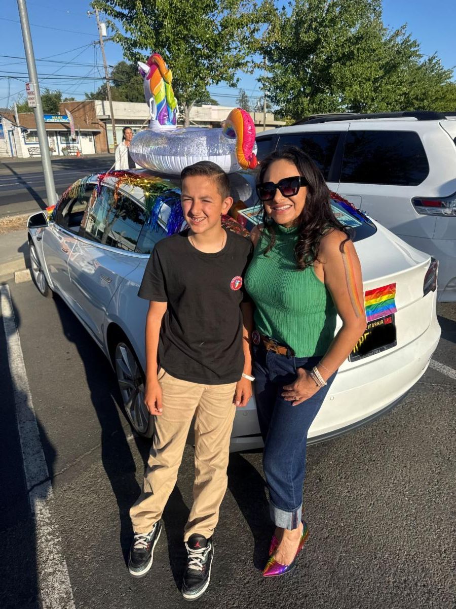 Photo of two people, one teenage boy and an adult female standing next to each other smiling and looking at the camera. Behind them is a parked white car with rainbow decorations on it. An inflated unicorn pool toy is on top of the car and rainbow streamers are hanging out the windows. 