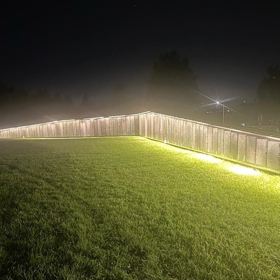 Photo is of the Wall the Heals illuminated and glowing at night. The wall is displayed across a very green lawn and the night sky is dark. 