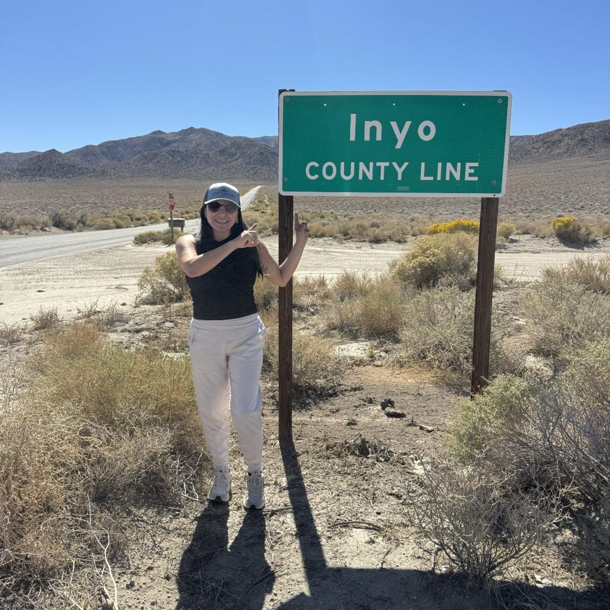 Photo is of a woman standing next to a green road sign that reads ‘Inyo County Line.’ The landscape is dry high dessert with lots of sagebrush and sand.