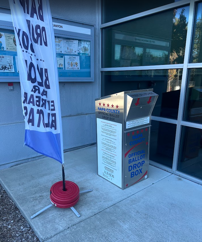 Photo depicts a large stainless steal official Napa County Ballot Drop box. Next to it is a tall flag that says ‘Ballot Drop Box’ on it. The drop box and flag are outside of American Canyon City Hall located at 4381 Broadway in American Canyon.  