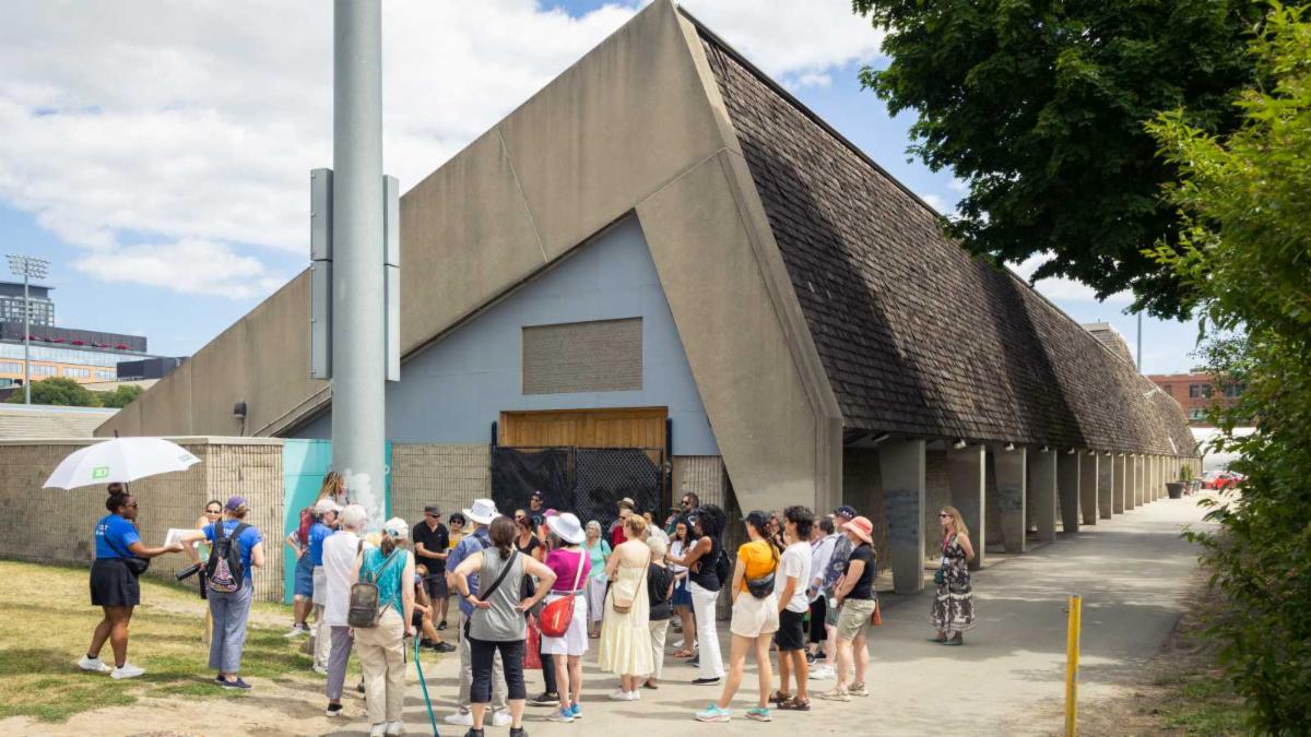 Group of people stand in front of a long triangular roofed building.