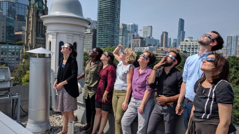 Eight individuals all wearing eclipse glasses look up at the sky. City view appears in the background and includes the St. James Cathedral bell tower and tree canopy of St. James Park.