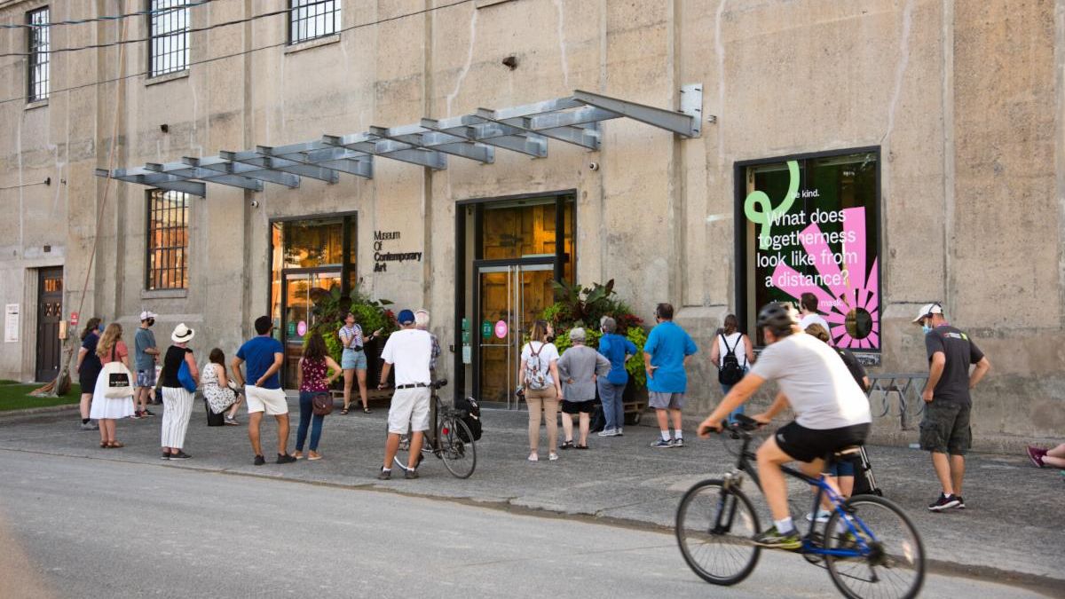 Group of people are gathered along a sidewalk at a building entrance. Sign at door reads Museum of Contemporary Art. A person cycles by on the road.
