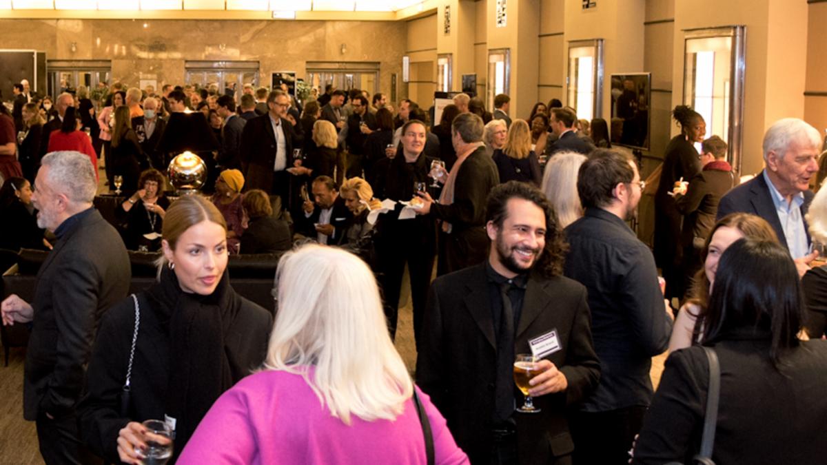View of crowd of people in a room at a reception with drink and food being held by many as they congregate in different groups and areas.
