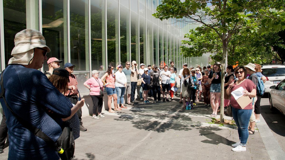 A woman in blue jeans and holding a microphone is talking to a large group of people who stand on the left and are lined up on the sidewalk in front of a metal and glass building.
