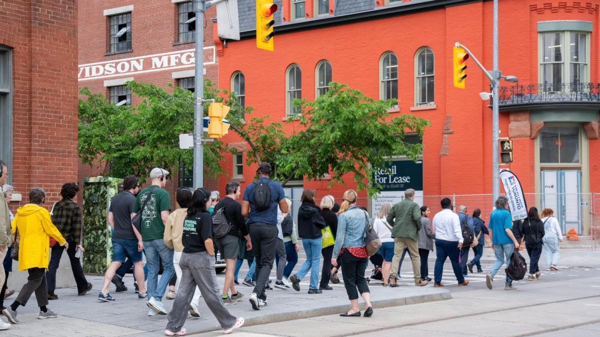 A large group of people walk along the sidewalk toward a three-storied and red-orange building.