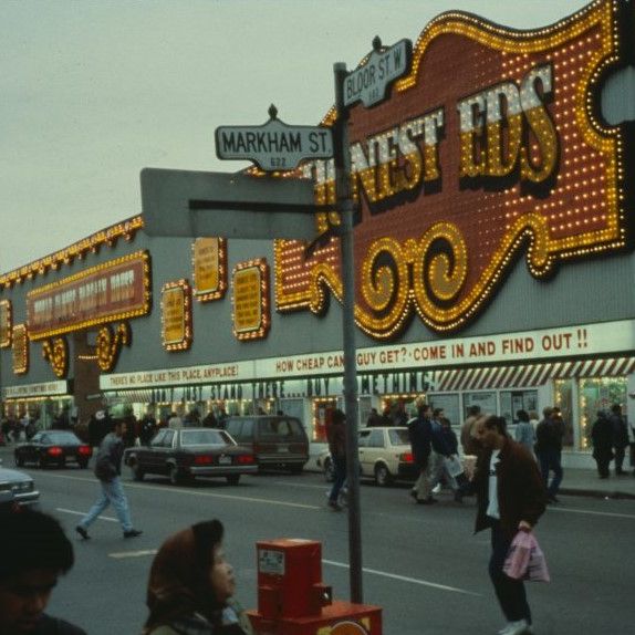 Street view of a large building with many bright neon signs in yellow and red. We can read Honest Eds on the largest one.