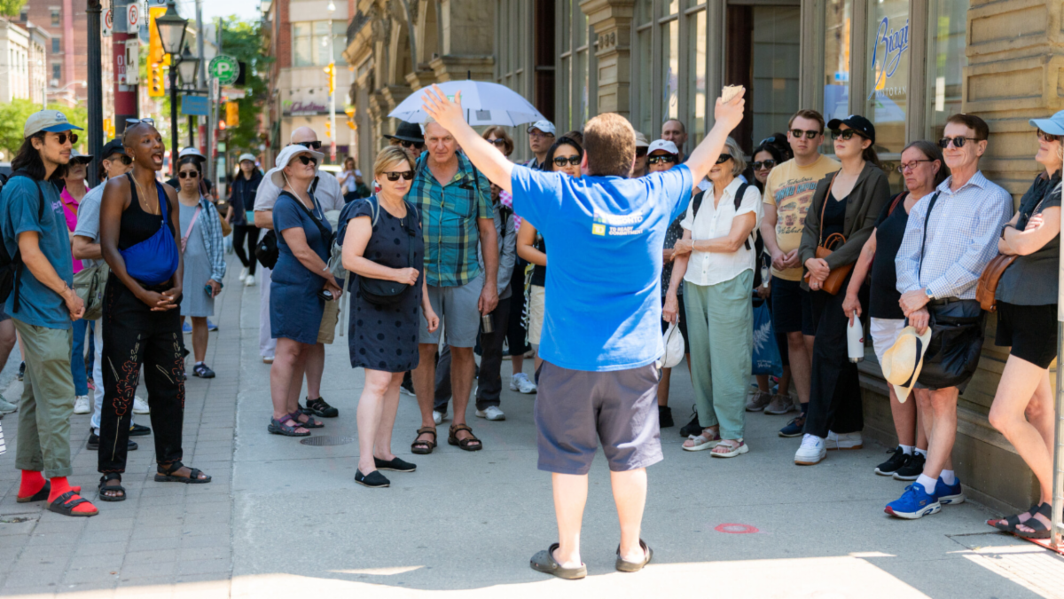 A group of people gather along a paved sidewalk and look toward a person dressed in blue and turned away from the camera. That person in the centre is gesturing with their arms up in the air.