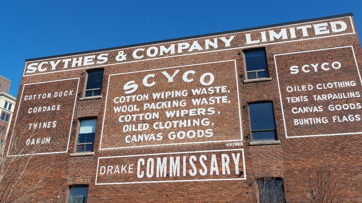 Photo of a red-brick building that has white writing painted on it. The writing reads, "Scythes & Company Limited. Scyco: Cotton wiping waste, wool packing waste, cotton wipers, oiled clothing, canvas goods / Drake Commissary." The sky above the building is bright blue.