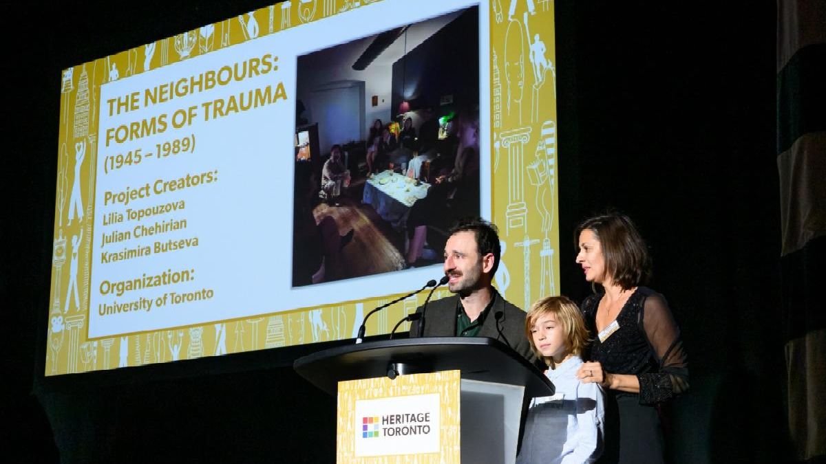 Two people and child at podium with one speaking into a microphone. Screen behind shows an image with text reading The Neighbours and names of project team members.