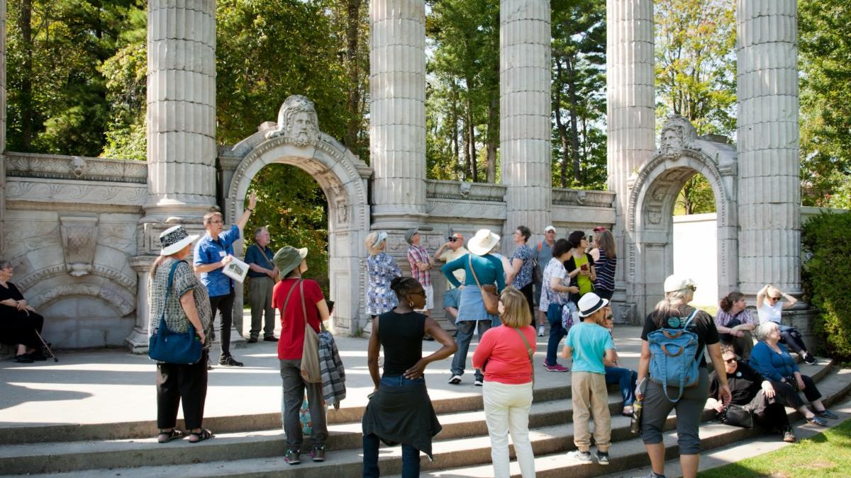 A group of people stand on a series of steps leading to a platform flanked in the background by an archway and a series of white columns.