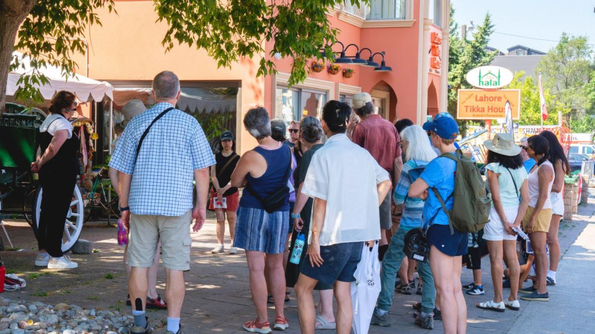 A group of people gather in front of a salmon pink building and listen to a speaker who is on the left of the image