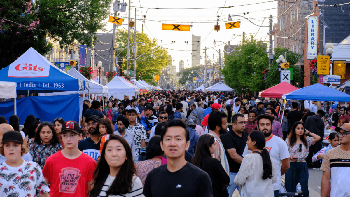Photo faces west down Gerrard St E. at the intersection of Gerrard St and Ashdale Ave. Tented booths with various vendors line both sides of the street and the road is filled with a large crowd of people visiting the festival.