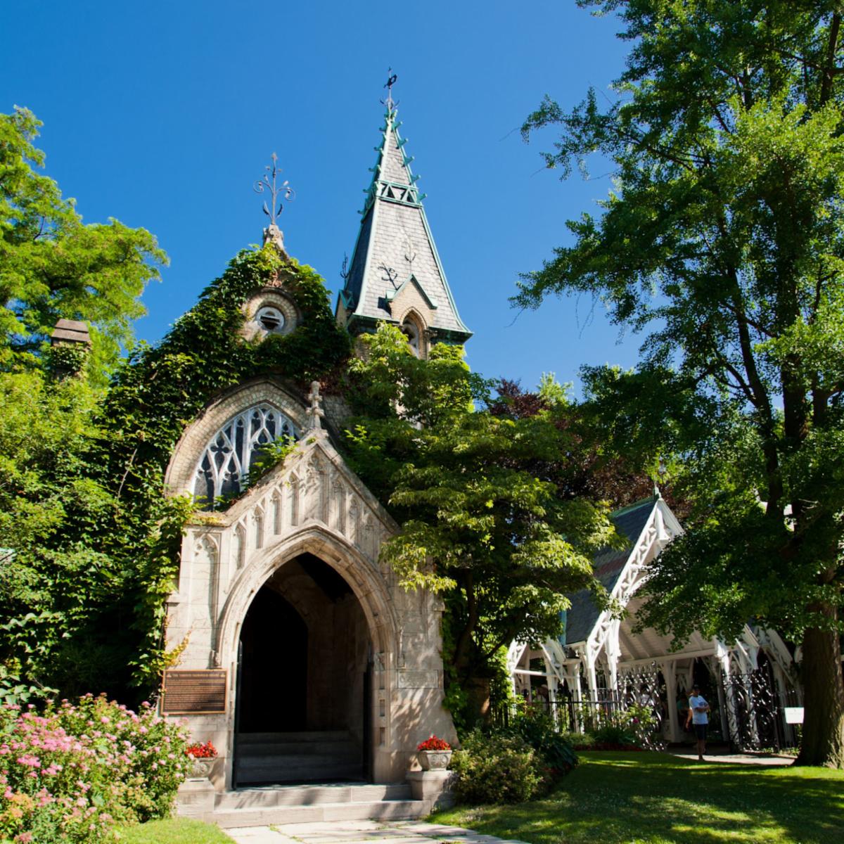 With a brilliant blue sky above, the photo centres on a Victorian gothic chapel, showing an arched entrance in the foreground and a stained window and spire in the background.
