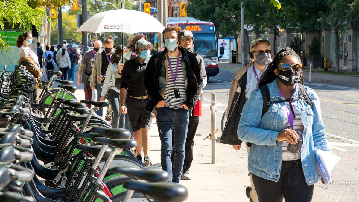 Woman wearing mask walks on a sidewalk next to a bike share station. Behind her a line of people follow.