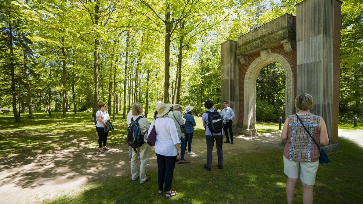 People are standing with their backs to the camera on a dirt path surrounded above by a canopy of trees in leaf looking at a stone and red brick arch.