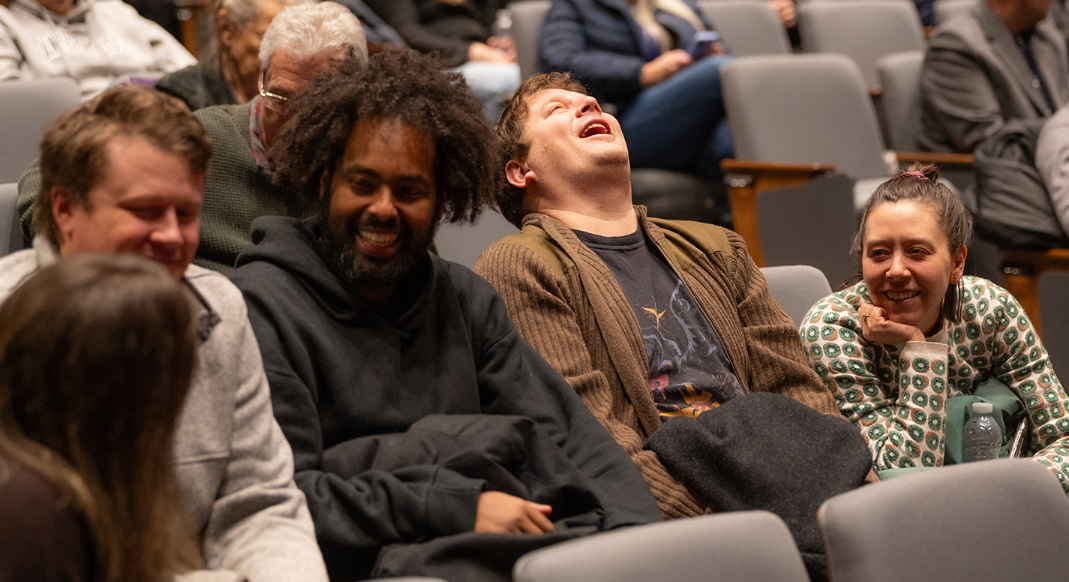 Close up of people sitting in auditorium chairs and laughing while conversing with each other. Five people can be seen interacting in the foreground with more people in the background partially seen.