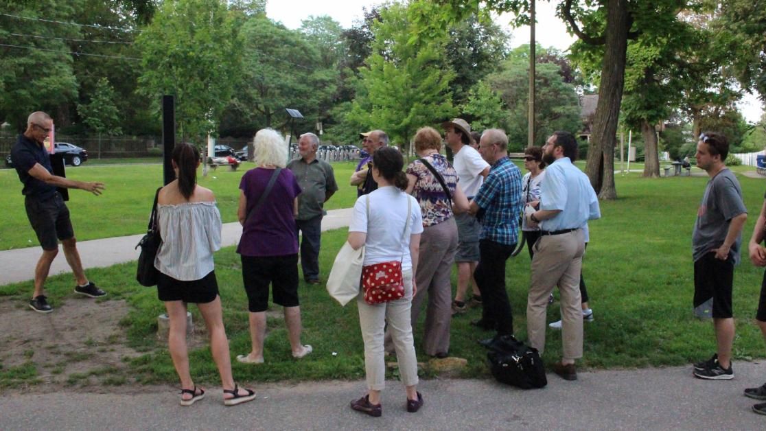 Group of people in park setting with backs to camera listen to speak on the left who gestures with his hand.