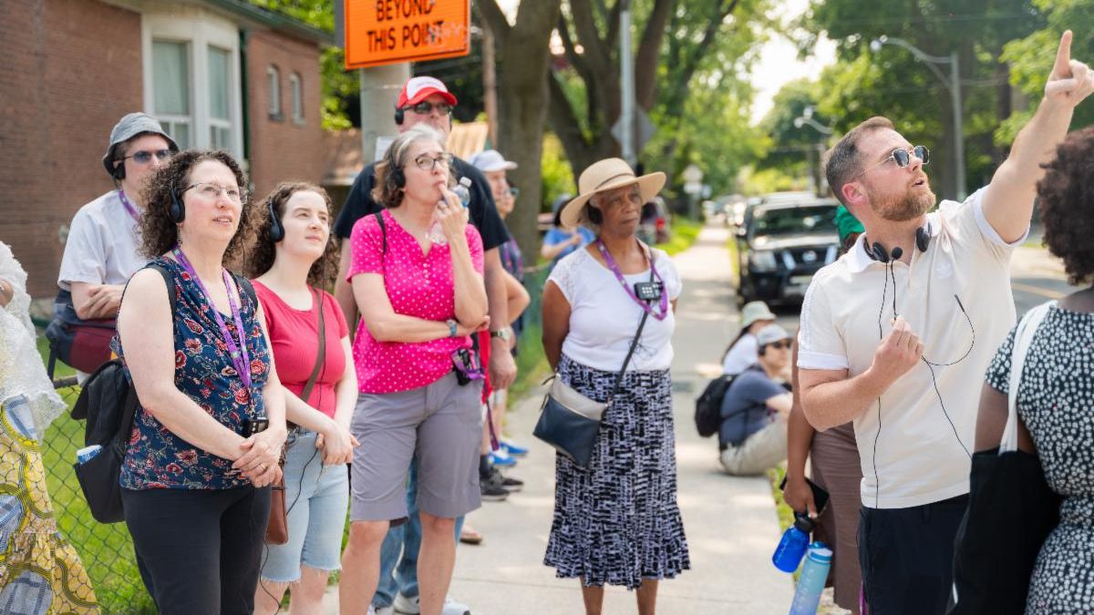 A group of people stand on a street corner in what looks like a residential neighbourhood. They are staring to the right and listening to a speaker who is pointing a finger to the top-right.