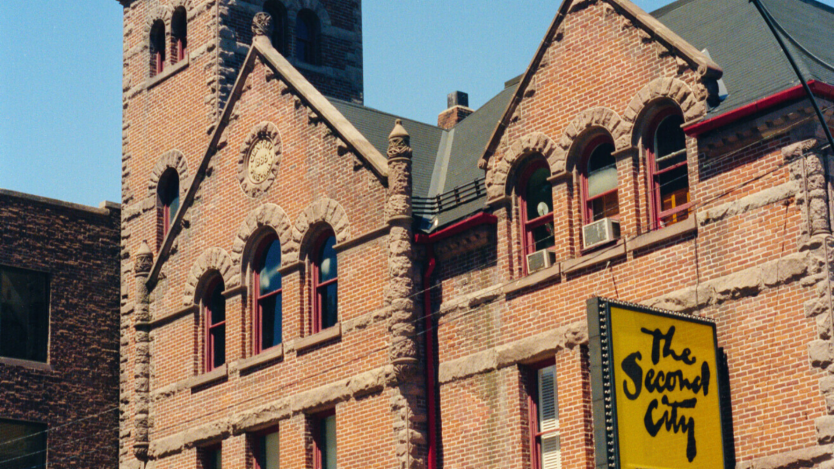 Photo of red brick building three-storeys tall with a tower. A yellow billboard advertises "Second City" in black lettering