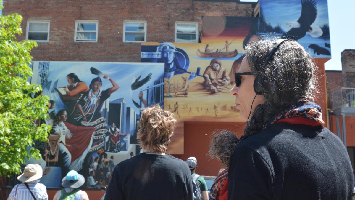 Two large colourful murals are in the background on a brick-wall building. People are in the foreground looking at the murals while the person to the far right is staring to the left. 