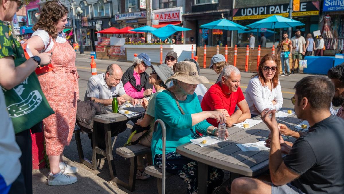 People stand and sit around and at tables next to a road. People seated are eating. Blue umbrellas and a streetscape are visible in the background.