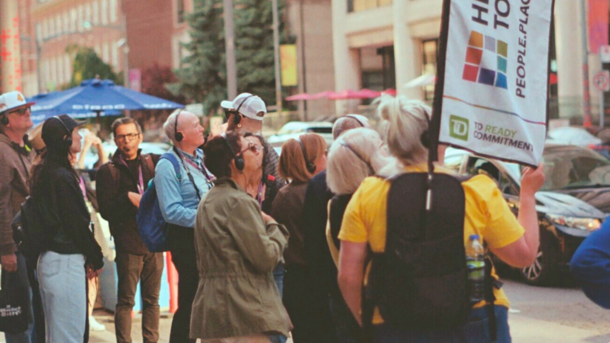 A tour group stands on a sidewalk next to a busy road lined with tall buildings. Members of the group are wearing audio headsets and looking across the street. A tour volunteer can be seen at the front of the image wearing a backpack with the Heritage Toronto flag.