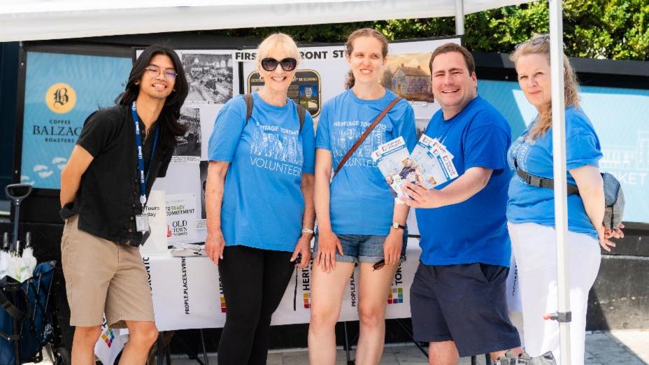 Three people wearing blue shirts, with one holding a Heritage Toronto flag, are seen walking and smiling.