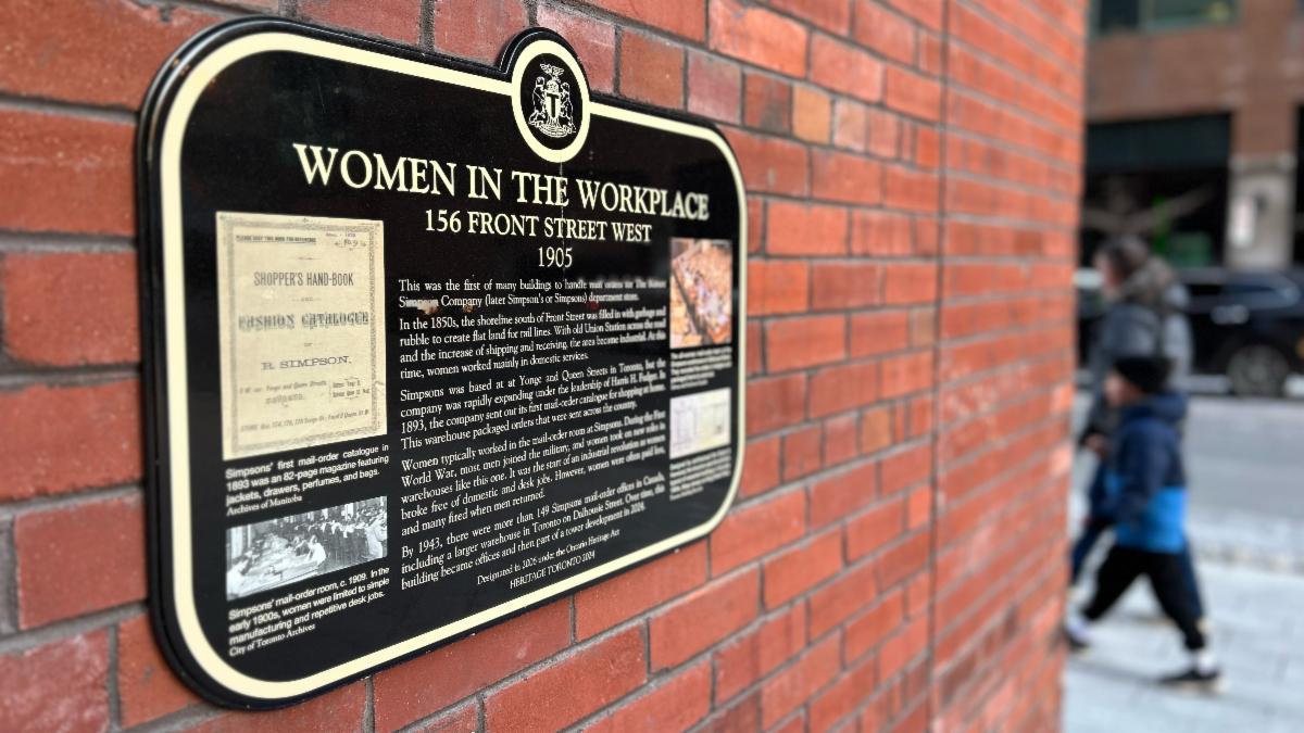 Image of a historical plaques attached to a red brick wall. In the background two people walk past.