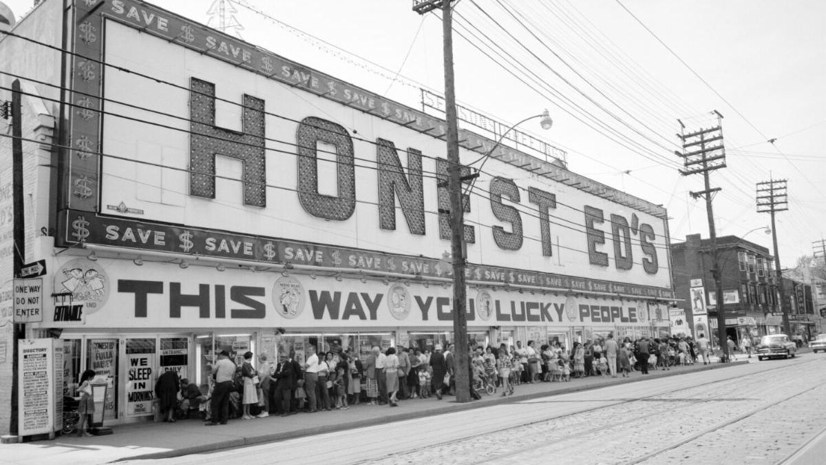 Street view of a large building with many bright neon signs in yellow and red. We can read Honest Eds on the largest one.
