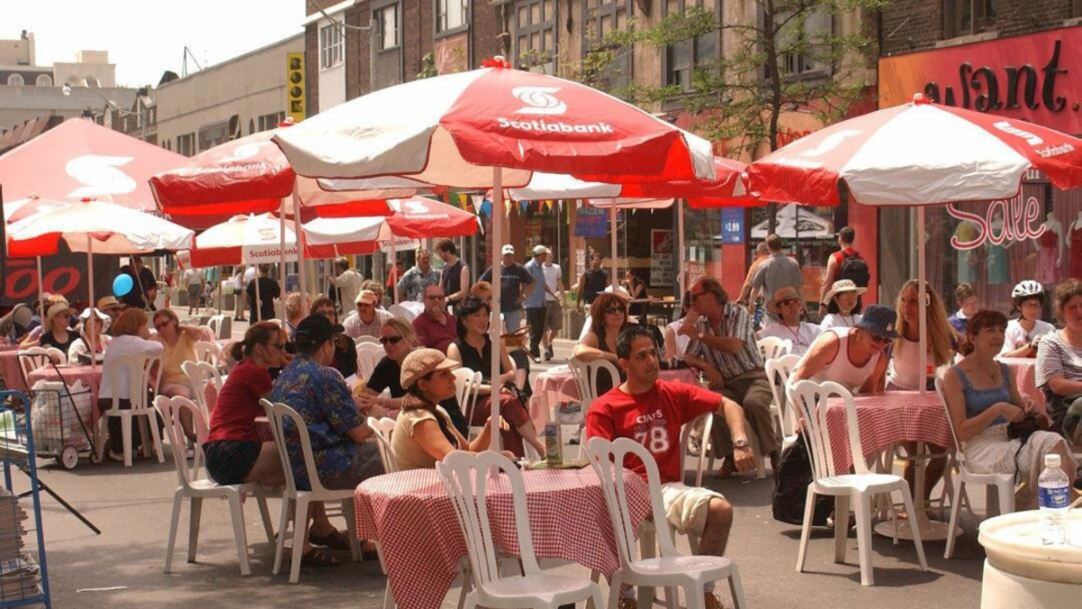 A crowd of people sit at round tables underneath red and white umbrellas.