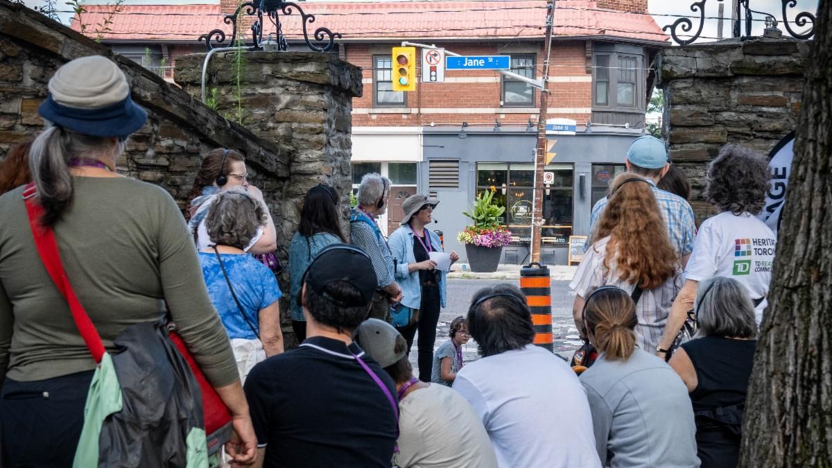 Group of people seated and standing with back to the camera listen to a person speaking at the front of the group.