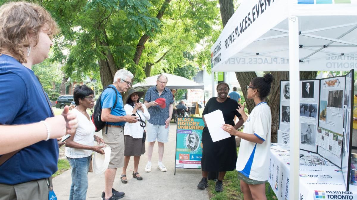 Two women speak in front of a table with a tri-fold display board on it under a tent next to a display sign advertising a walking tour. In front of them four people listen to them talk from the adjacent sidewalk.