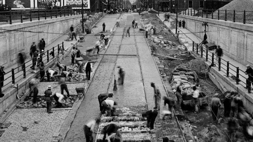 Black and white photograph of many workers building an underpass on an existing city road. In the distance are trees and buildings.